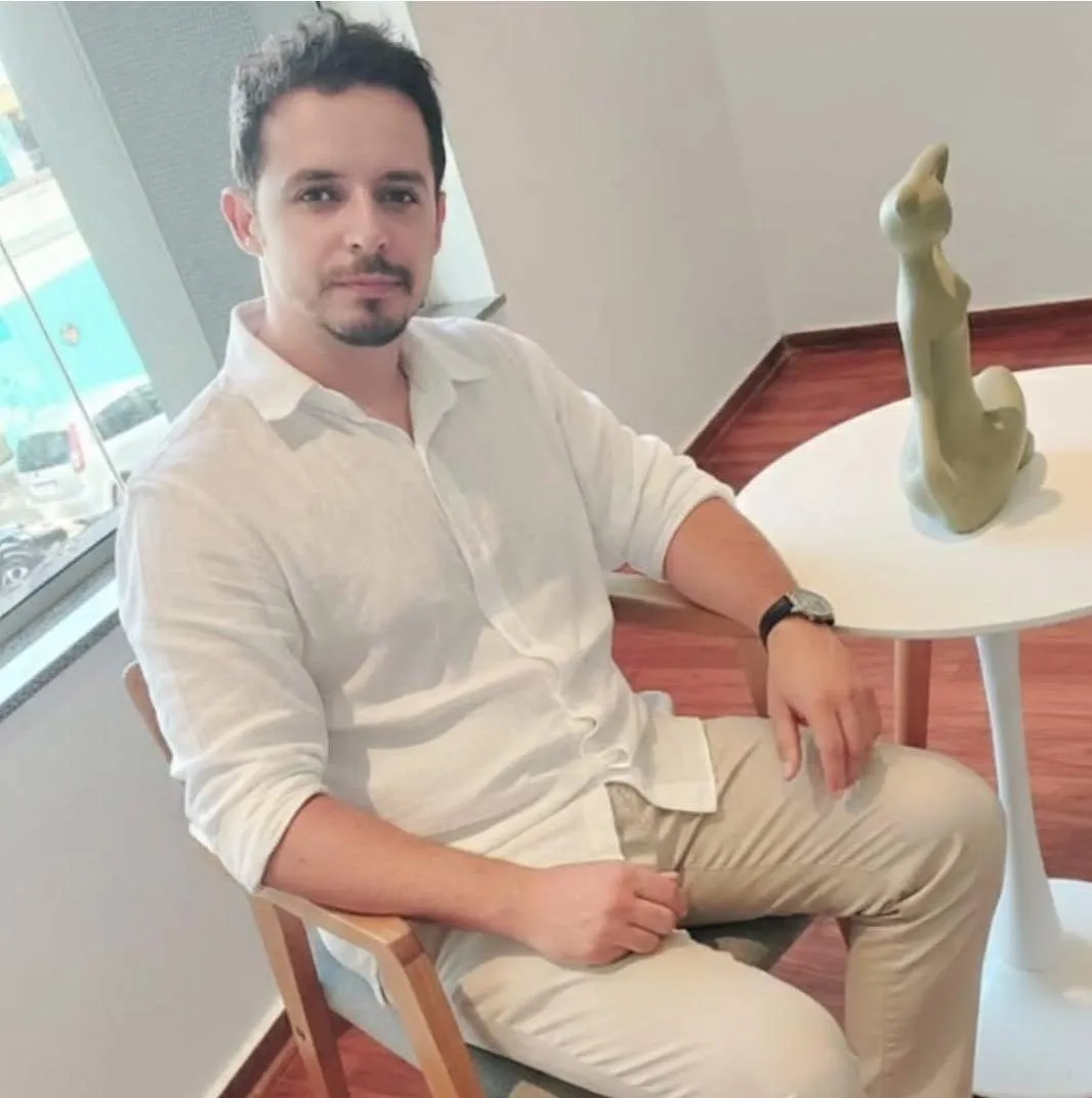 João Paulo de Souza Moreira, sentado perto de uma janela, de camisa branca
